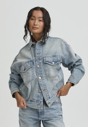 Kobieta o falowanych ciemnych włosach, ubrana w jasnoniebieską kurtkę dżinsową i pasujące dżinsy, pozująca z rękami w kieszeniach kurtki na jednolitym tle.