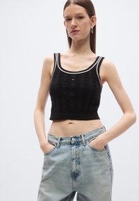 Juodas megztas trumpas viršus, pasižymintis ribuota tekstūra, plačiais petnešėlėmis ir kontrastingu baltu iškirptės apvadu, derinamas su šviesiai mėlynais aukšto liemens džinsais.