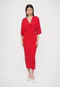 Sarkana kleita ar dziļu V-veida kakla izgriezumu un savelkamu detaļu viduklī, ar platām piedurknēm un pieguļošu siluetu. Gluds audums un vidējais teļa garums.