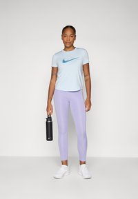 Světle modré tričko Nike s tyrkysovým logem, kombinované s levandulovými legínami. Žena drží černou láhev na vodu a má na sobě bílé tenisky.