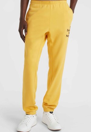 Geel sweats met een elastische tailleband en boorden, voorzien van zijzakken en zwarte bedrukte tekst op de linkse heup. Gecombineerd met witte sneakers.