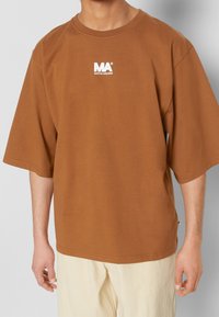 Camisa de algodón de gran tamaño en color marrón con mangas cortas, que presenta un logo blanco en el pecho. Fabricada en una suave mezcla de algodón. Textura lisa.