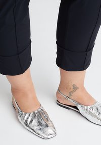 Füße tragen silberne, strukturierte Slingback-Ballerinas und schwarze gekürzte Hosen, rechts am Fußgelenk ist ein Tattoo sichtbar.