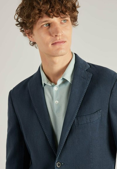 Marineblazer van gestructureerde stof, met een enkele knoopsluiting, gelapte revers, en een borstzak, gedragen over een lichtblauw poloshirt.