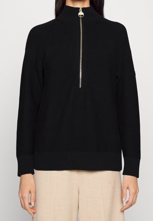 Femme portant un pull en maille noir à manches longues avec un col montant et une demi-fermeture éclair dorée, assorti à un pantalon beige sur fond uni.