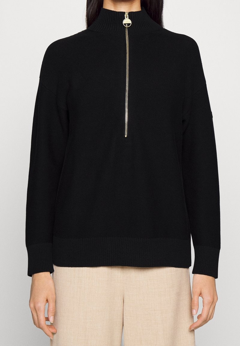 Femme portant un pull en maille noir à manches longues avec un col montant et une demi-fermeture éclair dorée, assorti à un pantalon beige sur fond uni.
