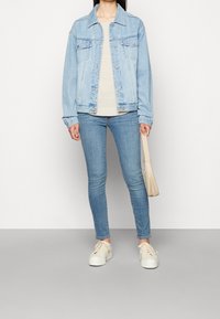 Giacca di jeans azzurro chiaro con due tasche frontali. Indossata sopra una maglia beige, abbinata a jeans skinny azzurro chiaro e sneakers bianche. Borsa beige in mano.