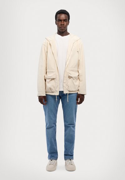 Svetlo bežjakna z kapuco z dvema sprednjima žepoma, z vrvico, oblečena čez belo majico, v kombinaciji z modrimi kavbojkami in svetlimi teniskami.