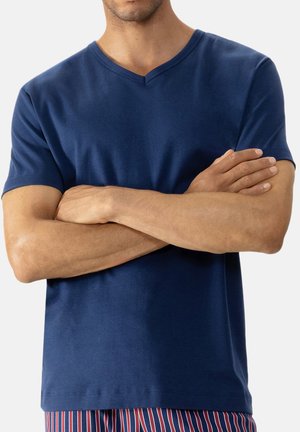 Mężczyzna w granatowej koszulce V-neck z założonymi rękami, widoczny od połowy klatki piersiowej do bioder, ubrany w paskowane czerwono-niebieskie spodnie.