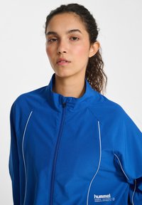 Blå zip-jacka gjord av texturerat material, med vita detaljer längs sömmarna och en hög krage, samt en logotyp på bröstet.