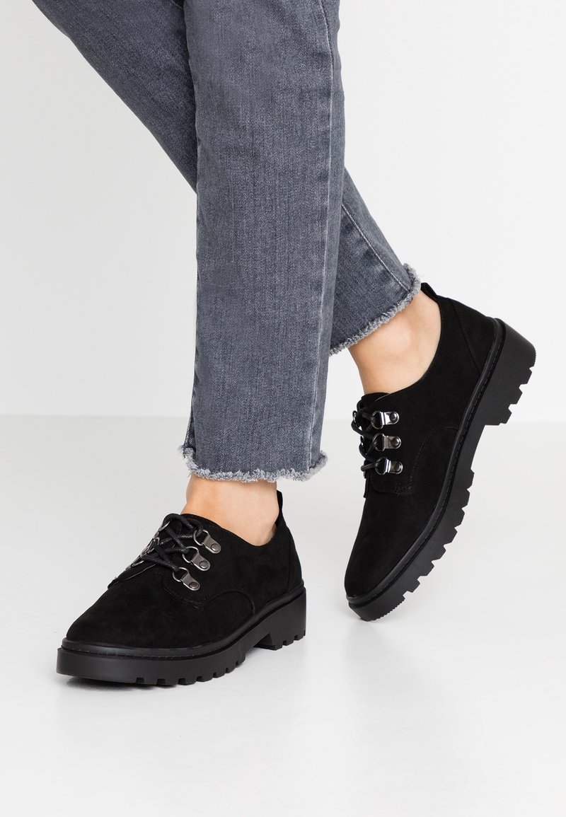 Chaussures en daim noir à bout rond, dotées de six œillets et de semelles en caoutchouc noir épais, associées à un jean gris avec un ourlet effiloché.