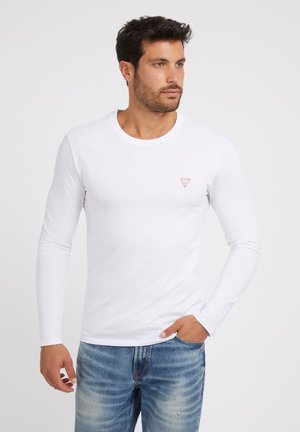 Weißes Langarm-T-Shirt aus Baumwolle mit einem kleinen roten dreieckigen Logo auf der Brust, kombiniert mit hellblauen Jeans.
