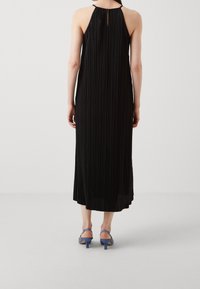 Czarna plisowana maxi sukienka z halterem i detalem w kształcie dziurki z tyłu, w zestawieniu z niebieskimi sandałami na obcasie. Gładka tekstura materiału.