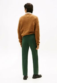 Brązowy, teksturowany sweter z dekoltem w serek i prążkowanymi mankietami, w połączeniu z prostymi zielonymi spodniami. Czarne buty dopełniają zestaw.