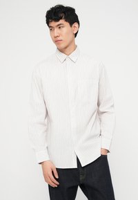 Νέος άνδρας με μαύρα, σγουρά μαλλιά, φορώντας λευκό πουκάμισο με μακριά μανίκια και λεπτές κάθετες ρίγες, καθώς και σκούρο τζιν, στέκεται με το ένα χέρι στην τσέπη.