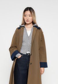 Woman wearing a brown trench coat with navy cuffs over a gray zip-up hoodie and dark blue jeans, standing with one hand in pocket against white background.