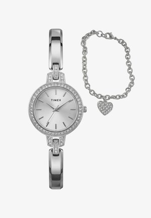 Srebrni Timex sat s okvirom ukrašenim kristalima i trakama u obliku šipki, uparen sa srebrnom narukvicom s lancem koja ima privjesak u obliku srca od kristala.