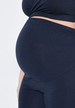 Schwangere Person, die dunkelblaue Umstandsleggings und ein passendes Oberteil trägt, wobei der runde Bauch und der dehnbare Bund betont werden.