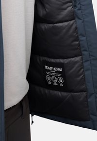 Veste isolante bleu marine avec une finition mate, dotée d'une doublure intérieure noire et d'une étiquette indiquant des propriétés de respirabilité et de séchage rapide.