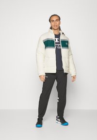 Puffjakke i creme med grønne og hvide striber, parret med en marineblå t-shirt og sorte bukser med refleksdetaljer samt sneakers.