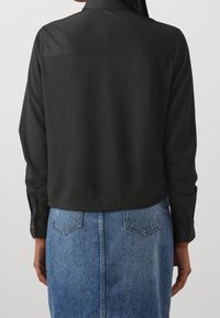Schwarzes, geripptes langärmeliges Hemd mit Kragen und geknöpften Manschetten, cropped Design, getragen über einem blauen Jeansrock mit aufgesetzten Taschen.