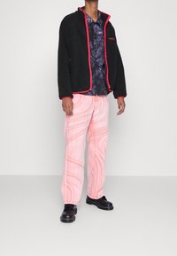 Černá fleecová bunda s červeným lemem, spojená s růžovými kalhotami se spirálovými vzory. Podpořena černými botami, vystavená na neutrálním pozadí.