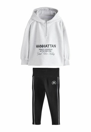 Sweatshirt à capuche gris avec zip, arborant un texte noir et une poche kangourou ; assorti à un legging noir avec des rayures blanches sur les côtés et un logo.