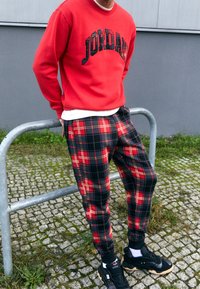 Man in een rode Jordan-sweatshirt, rode en zwarte geruite broek en zwarte sneakers, leunt tegen een metalen reling op een kasseien pad.