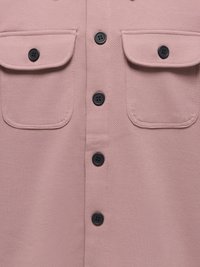 Gros plan sur un tissu rose poudré avec des boutons noirs à quatre trous et deux poches à rabat avec boutons sur un vêtement.