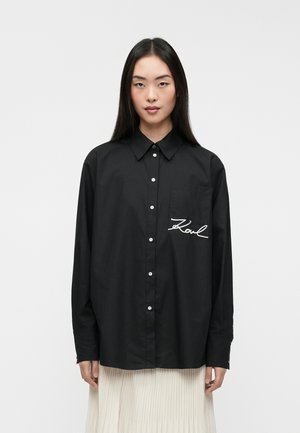 Must must shirt, mille taskuga ees, millel on valge tikandiga tekst "Karl." Valmistatud siledast kangast ja pikal varrukal.