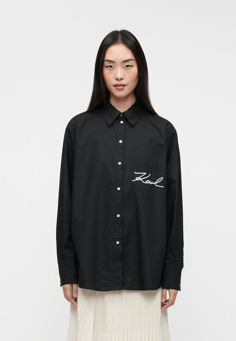 Camisa preta de botões com um bolso frontal, apresentando texto bordado em branco "Karl." Feita de tecido suave com mangas longas.