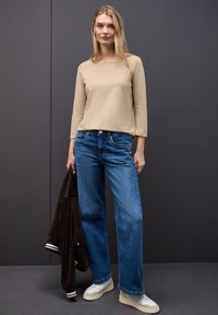 Maglietta beige a maniche lunghe, jeans blu a gamba larga, sneakers chiare, con una giacca marrone in mano. Tessuti morbidi, design casual e vestibilità rilassata.