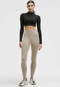 Top crop lungo nero a maniche lunghe abbinato a leggings beige a vita alta, entrambi realizzati in un materiale elastico. La modella indossa sneakers bianche.