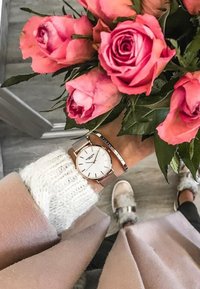 Reloj de pulsera rosa dorado con esfera blanca, correa de malla y combinado con una pulsera metálica. Mano sosteniendo un ramo de rosas rosas, suéter suave.
