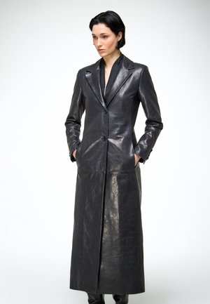 Kobieta z krótkimi czarnymi włosami, ubrana w długi czarny skórzany płaszcz z guzikami, z rękami w kieszeniach, na jednolitym białym tle.