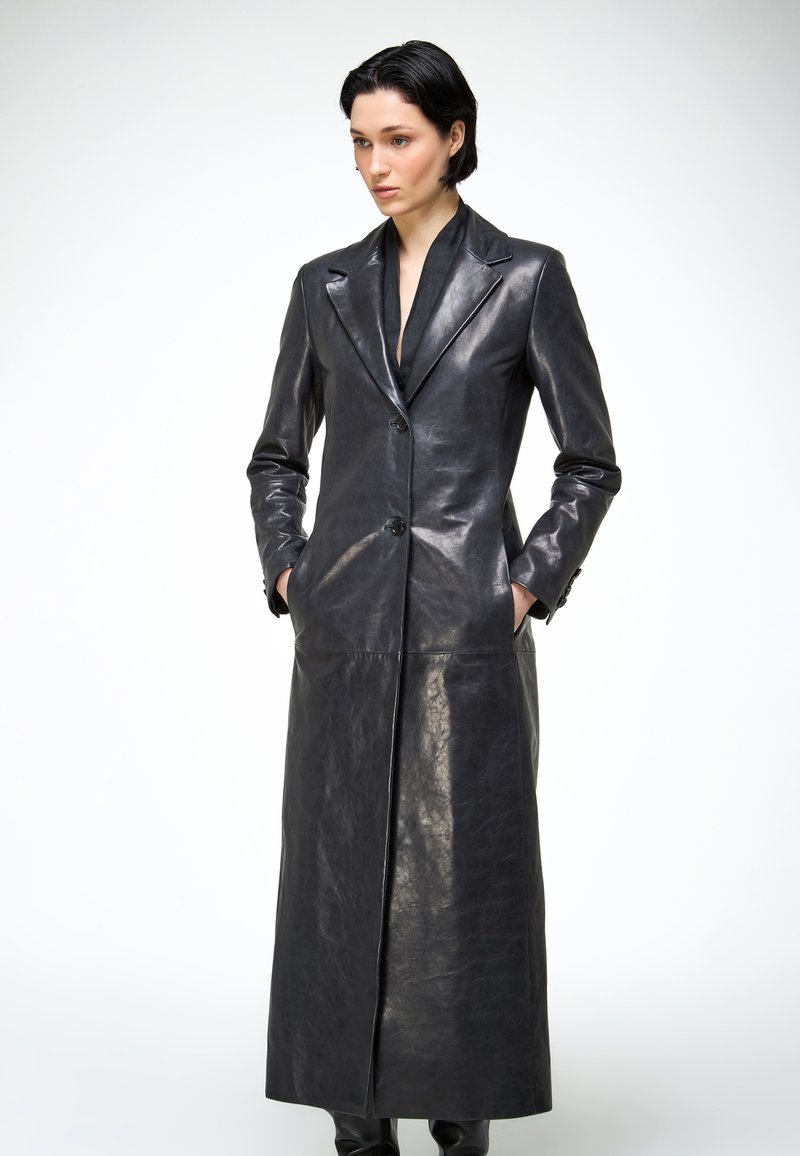 Femme aux cheveux noirs courts portant un long manteau en cuir noir à boutons, les mains dans les poches, sur un fond blanc uni.