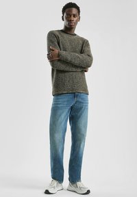 Gebreide trui in donkergroene en gouden tinten, ronde halslijn, gecombineerd met lichtblauwe rechtpassende jeans en grijze sneakers.