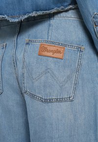 Light blue denim jeans feature a rear pocket with a brown leather label embossed with "Wrangler" and zigzag stitching details.