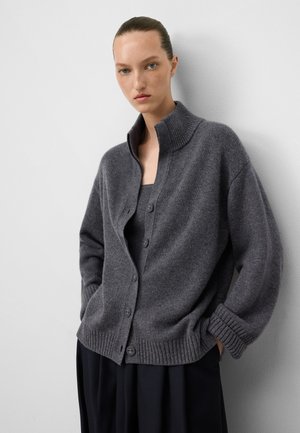 Vrouw in een grijze knopen cardigan met geribde kraag en boorden over een bijpassende top, staand tegen een witte muur met de handen in de zakken.