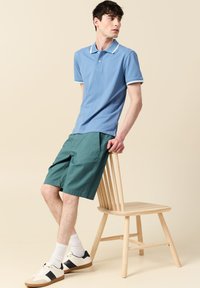 Vaaleansininen polo paita valkoisilla yksityiskohdilla, yhdistettynä vihreisiin shortseihin. Mallilla on valkoiset tennarit tummansinisin yksityiskohdin ja valkoiset sukat, istuen puutuolilla.