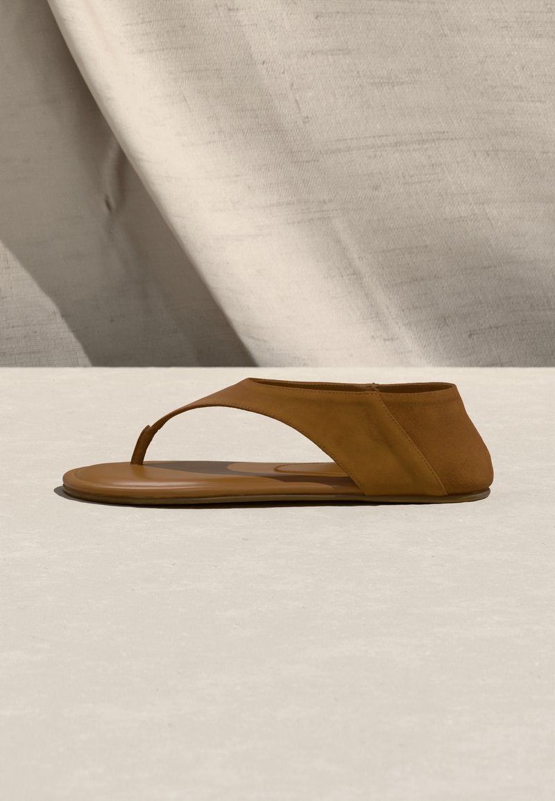 Sandale plate marron avec une large bride sur le dessus du pied et une lanière entre les orteils, posée sur une surface neutre avec un fond en tissu.