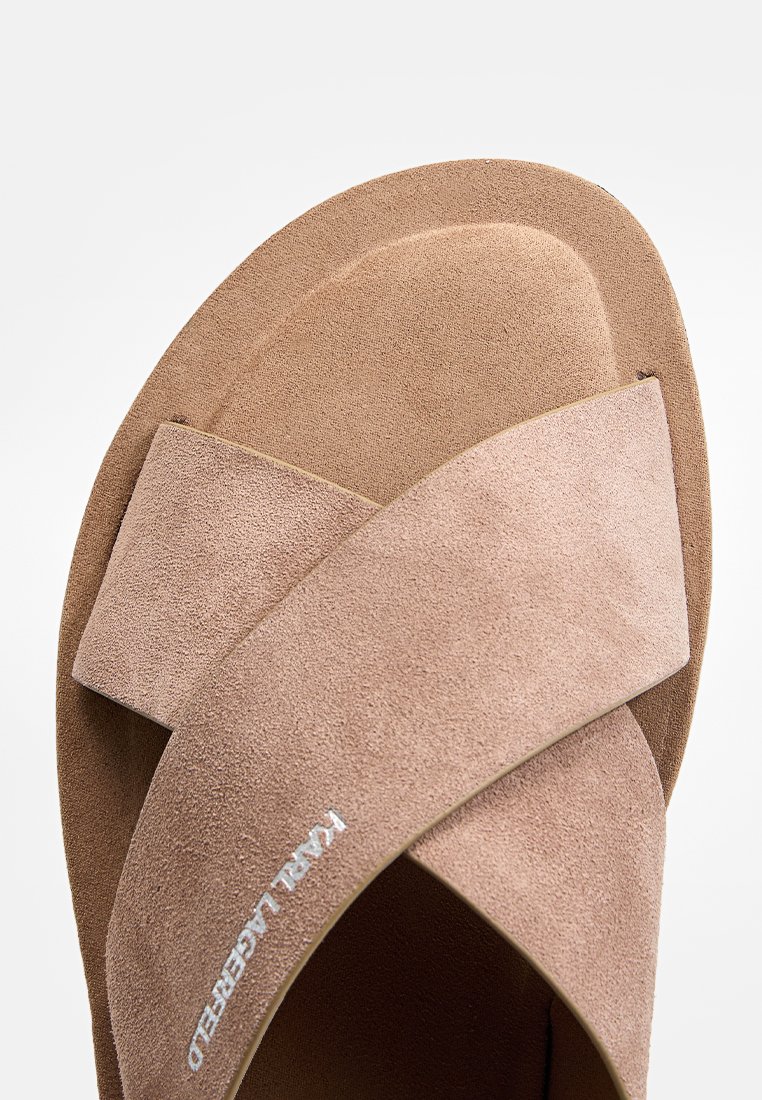 Braune Wildleder-Sandale mit Kreuzriemen, präsentiert sich mit einer strukturierten Oberfläche, einem gepolsterten Fußbett und einem weißen Logodesign. Glatte Sohle darunter.
