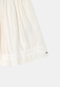 Valkoinen kerätty hameen helma, jossa on brodeerattu Tommy Hilfiger -teksti ja pieni metallinen Tommy Hilfiger -lipputunnus oikeassa alareunassa.