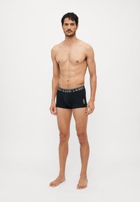Black boxer shorts made of soft fabric, featuring a logo waistband and a small embroidered design on the front. Model stands barefoot.