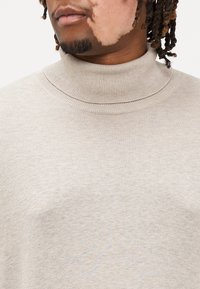 Pull beige à col roulé avec une texture côtelée, présentant un col plié et une silhouette ajustée. Le tissu semble doux et tricoté.