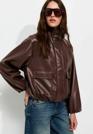 Vrouw draagt een oversized donkerbruine leren jas met grote frontzakken, blauwe denim jeans en grote donkere zonnebril tegen een effen achtergrond.