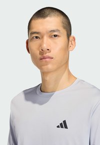 Mladý muž s krátkymi vlasmi v svetlosivom športovom tričku Adidas, čelí vpred s neutrálnym výrazom a jednoduchým pozadím.