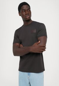 Mannlig modell med mørk hud iført mørkegrå EA7 T-skjorte og lyseblå jeans, poserer med armene krysset mot hvit bakgrunn.