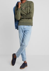 Sudadera verde oliva con logo impreso, jeans azul claro y zapatos gris oscuro. La sudadera tiene puños y dobladillo acanalados. Chaqueta de mezclilla colgada del brazo.