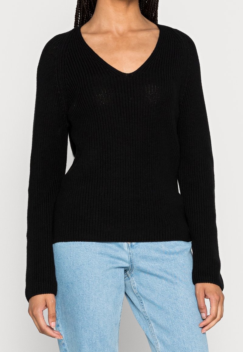Femme portant un pull noir en maille à col en V et un jean bleu clair, debout les mains détendues le long du corps devant un fond uni.